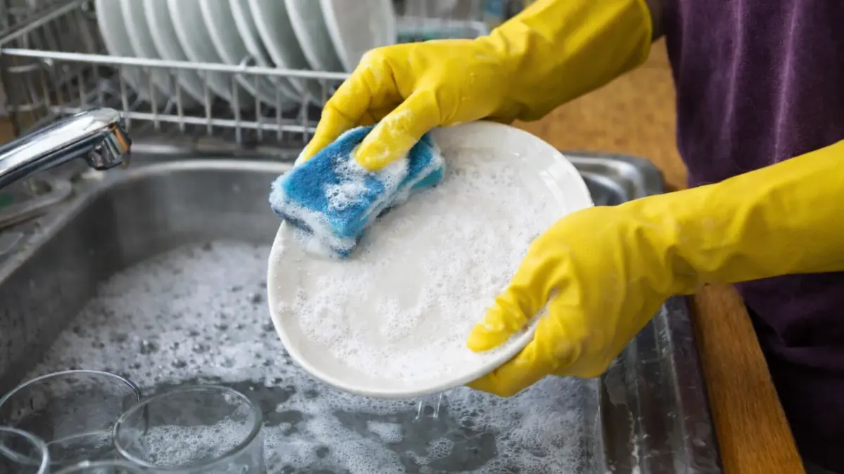عامل يقوم بغسل الأواني في حوض المطبخ مع قفازات صفراء وإسفنجة وصحن أبيض مع ظهور علم أستراليا في الخلفية ضمن سياق العمل في أستراليا بدون خبرة في وظيفة عامل غسل الأواني