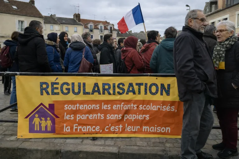 صورة واقعية لوقفة احتجاجية سلمية في بلدة فرنسية، يظهر فيها مهاجرون من أصول شمال إفريقية وشرق أوسطية خلف لافتة ملونة تطالب بتسوية أوضاع المهاجرين بدون أوراق في فرنسا، مع وجود علم فرنسا وأجواء شتوية طبيعية